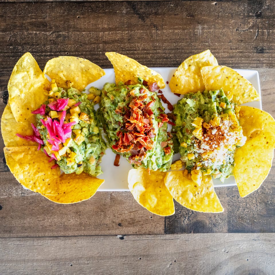 Three loaded guacamole servings topped with corn, pickled onions, bacon, and cotija cheese, surrounded by tortilla chips on a white platter.
