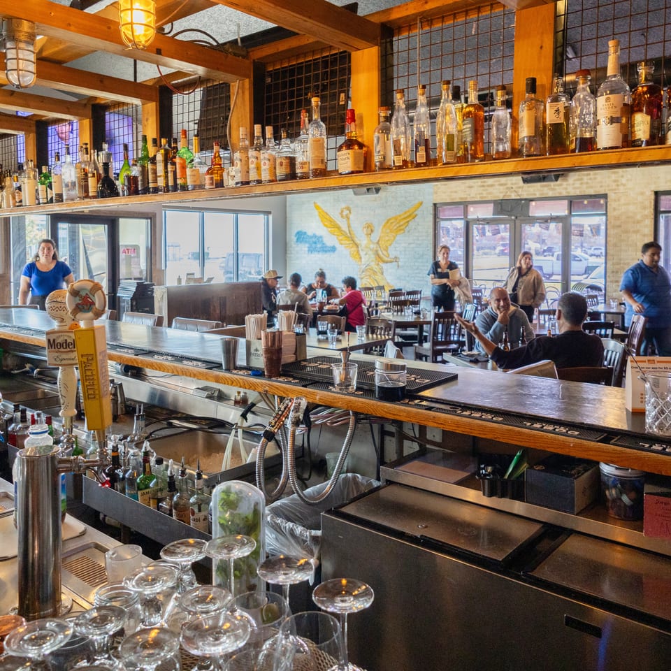 Bustling restaurant bar lined with liquor bottles, beer taps, hanging lights, tables, and patrons beneath a winged mural.