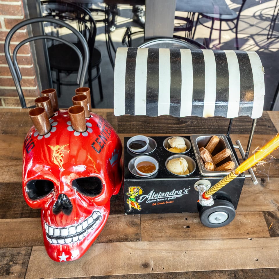 Colorful dessert cart displays churros, dipping sauces, ice cream, and copper cups beside a red skull-shaped container with a lit sparkler.
