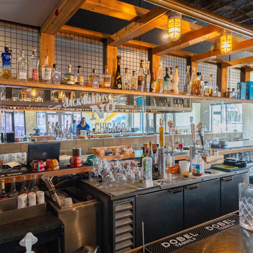Warmly lit restaurant bar lined with liquor bottles, hanging lanterns, taps, glassware, and a Jack Daniel’s No. 7 Chicago mirror.