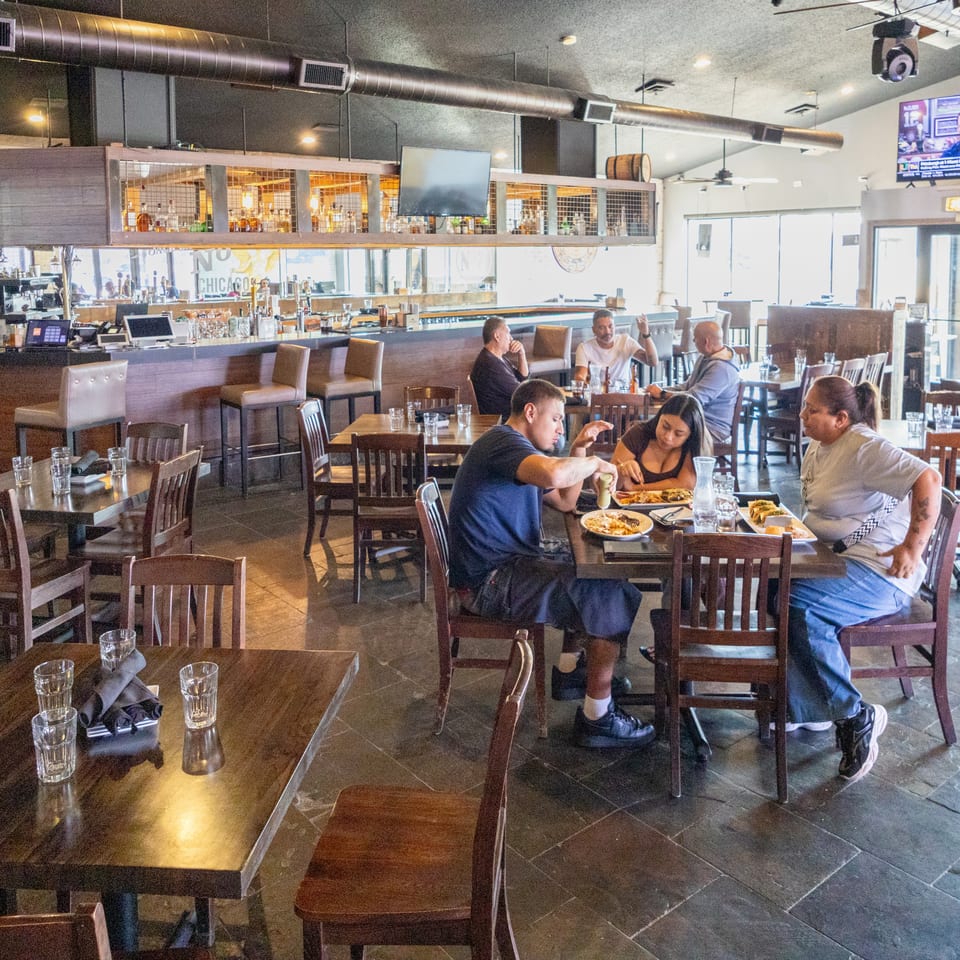 Bright casual restaurant dining room with bar, televisions, and guests enjoying meals at wooden tables.
