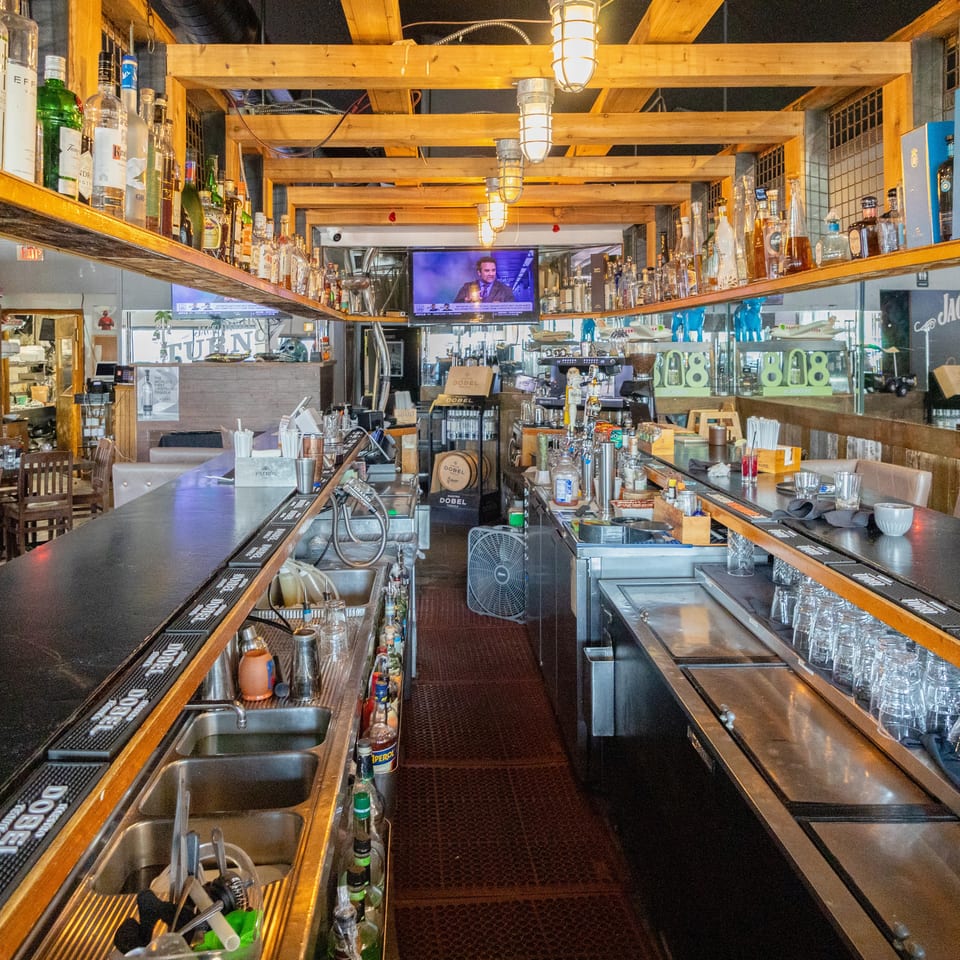Interior view of a well-stocked bar with liquor shelves, polished counters, taps, glassware, and overhead wooden beams.