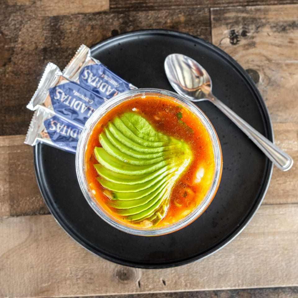 Bowl of orange soup topped with fan-sliced avocado, served on a black plate with spoons and condiment packets.