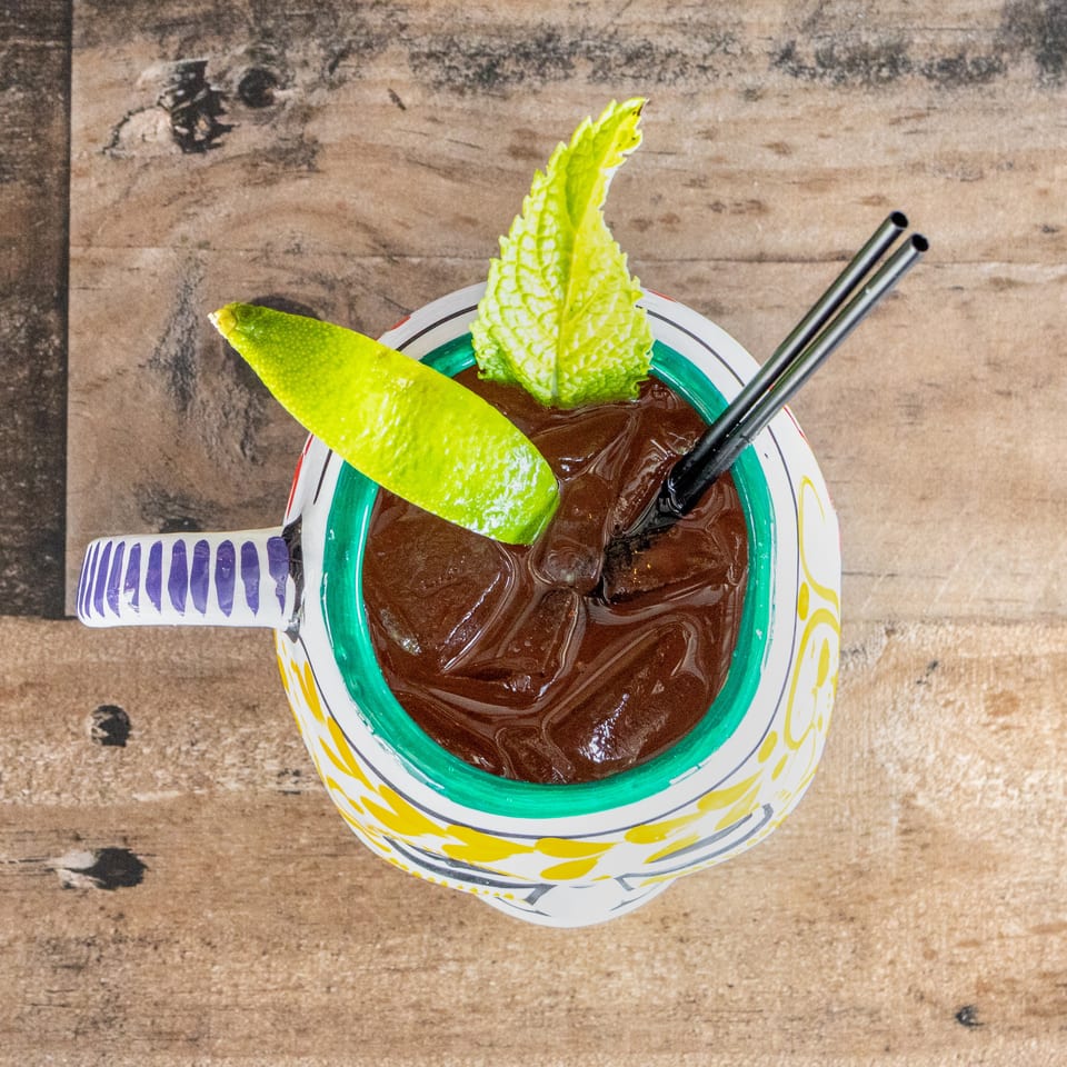 Iced dark cocktail garnished with lime and mint in a colorful patterned glass on a rustic wooden table.
