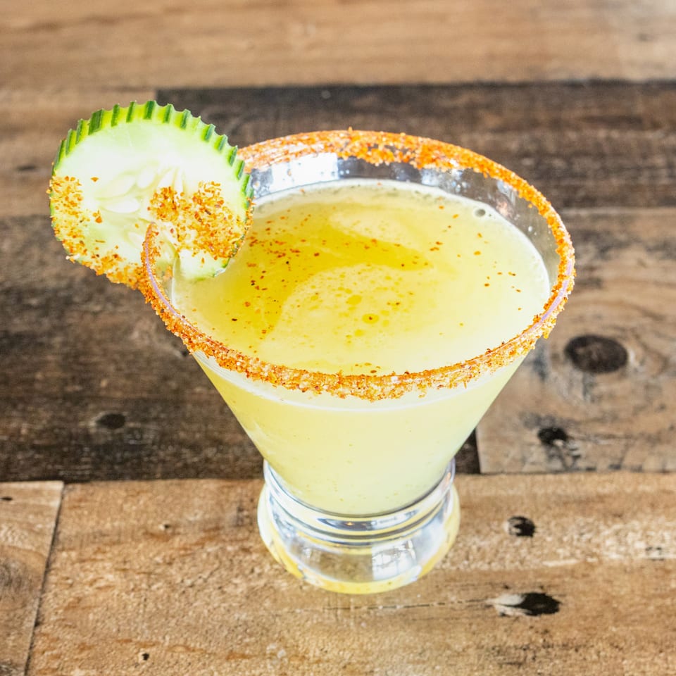 Pale yellow cocktail in a chili-rimmed glass, garnished with a cucumber slice, sits on a rustic wooden table.