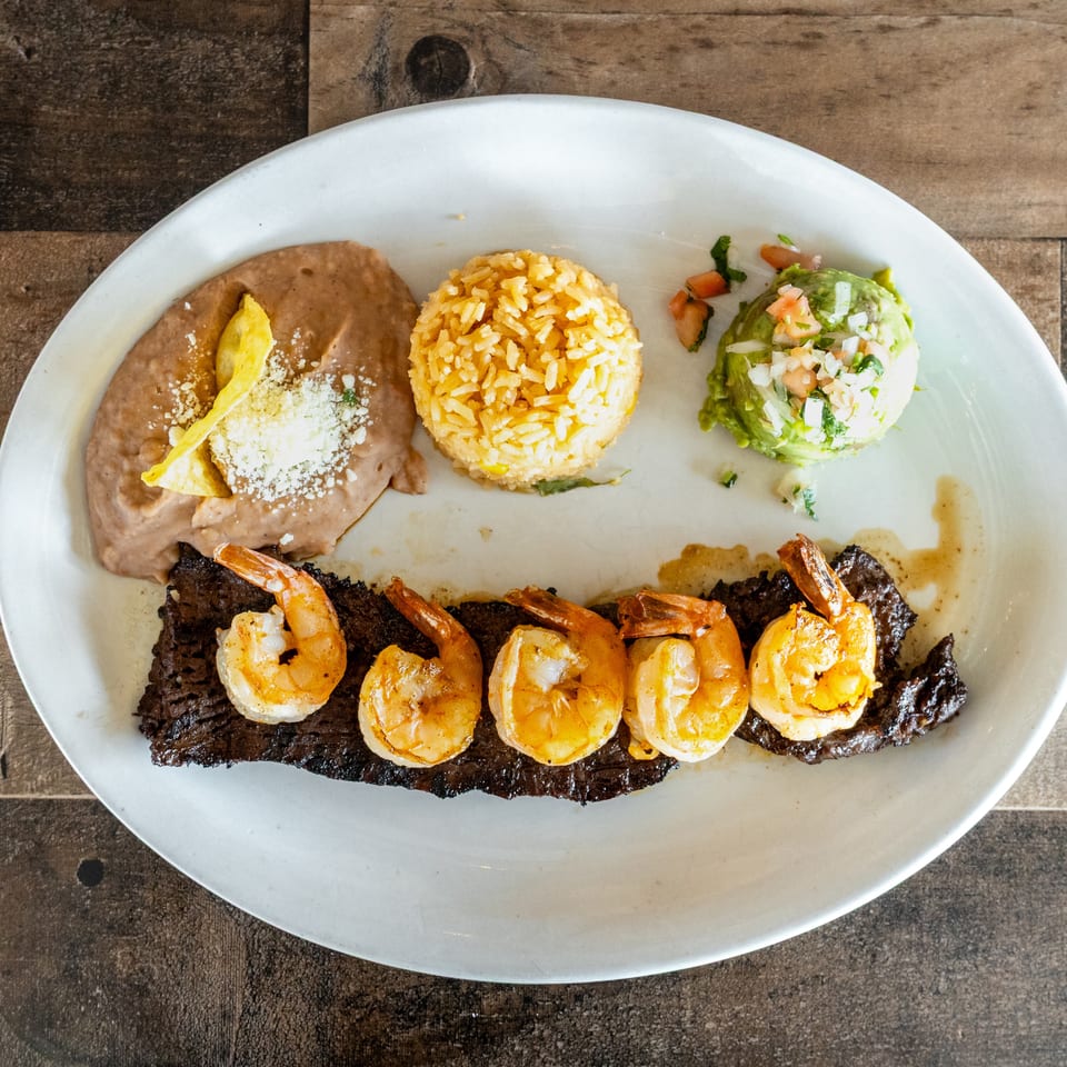 Plated Latin American meal featuring grilled steak topped with shrimp, rice, refried beans, guacamole, and tortilla chips.