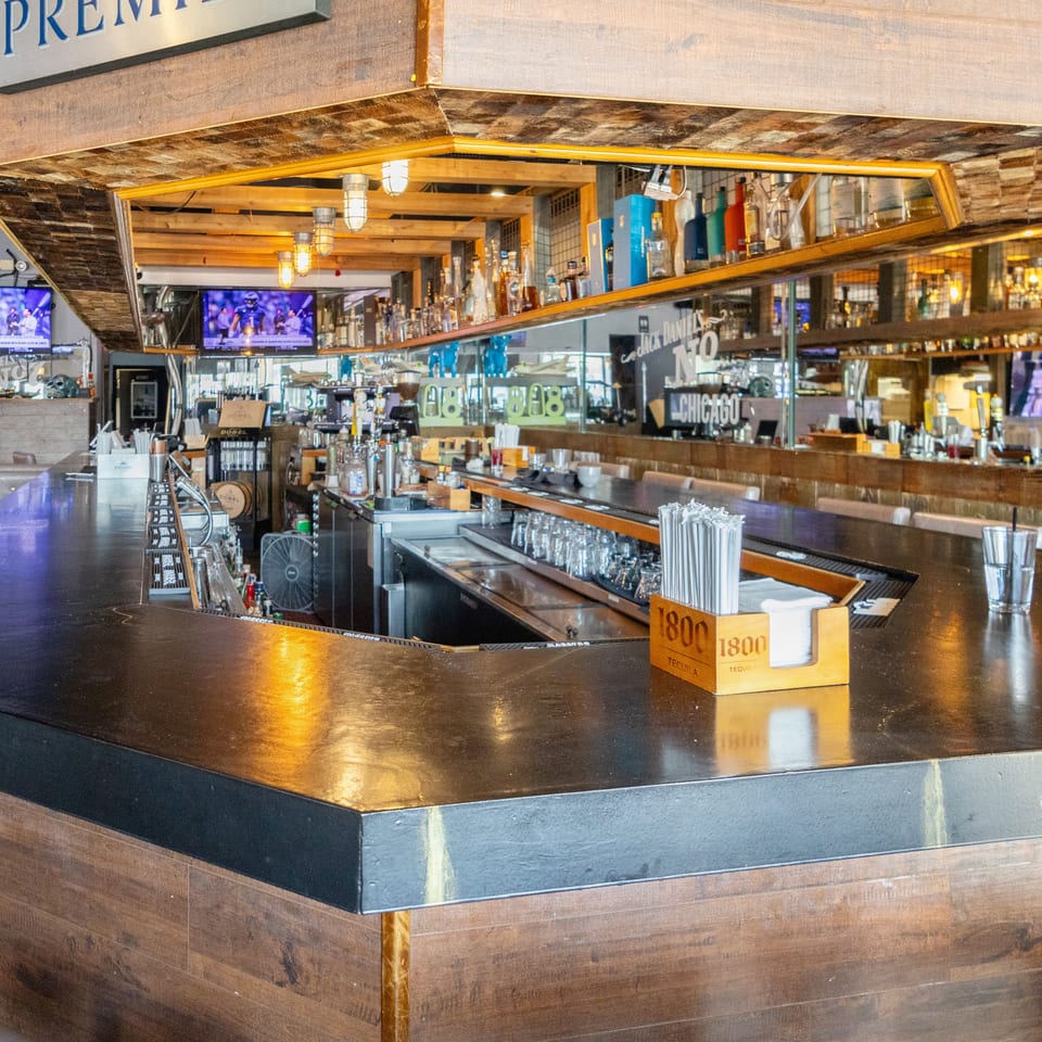 Spacious restaurant bar with polished counters, liquor shelves, mirrors, televisions, and warm wood-and-brick decor.