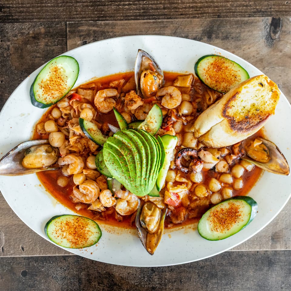 Seafood stew with shrimp, mussels, octopus, avocado, cucumber, and toasted bread served on a white platter.