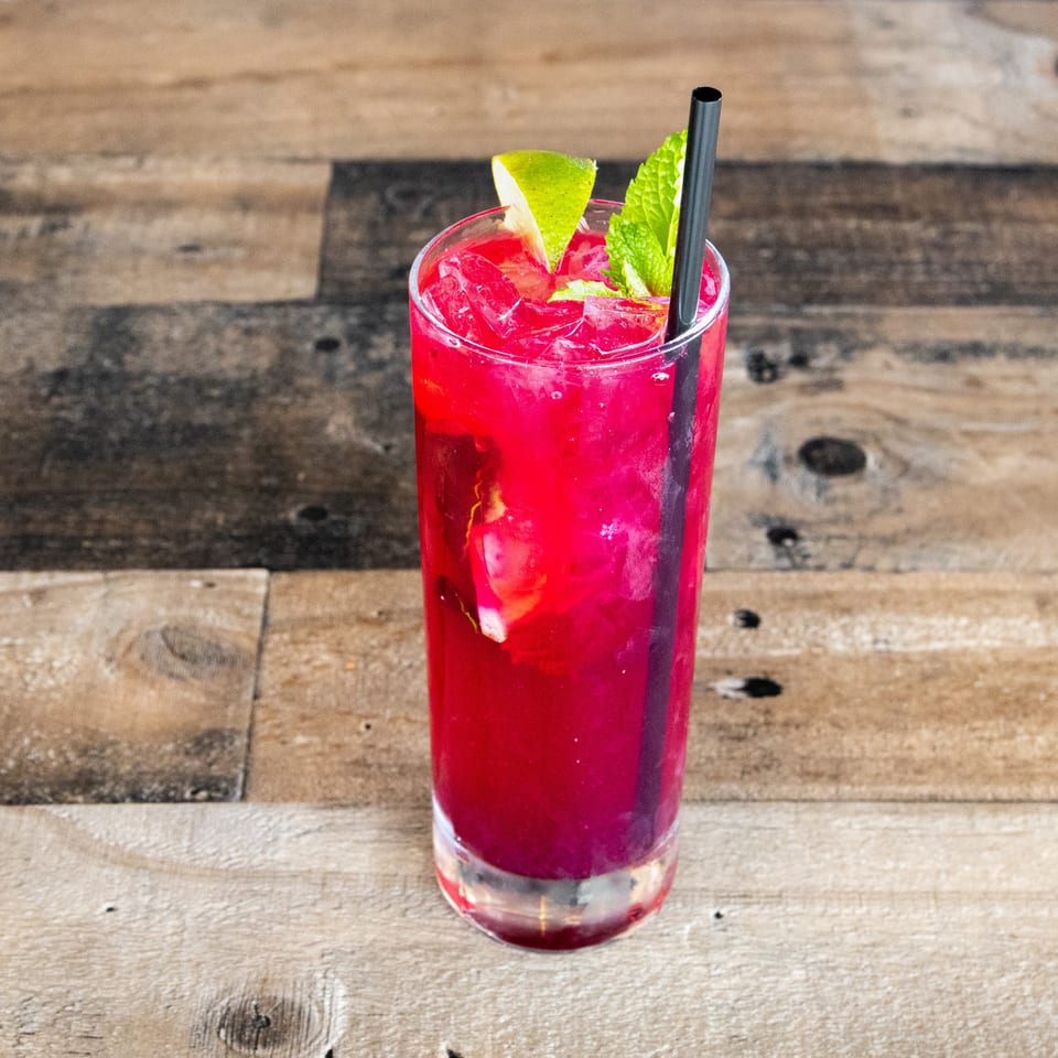 Bright red iced cocktail garnished with lime and mint, served with a black straw in a tall glass on a rustic wooden table.