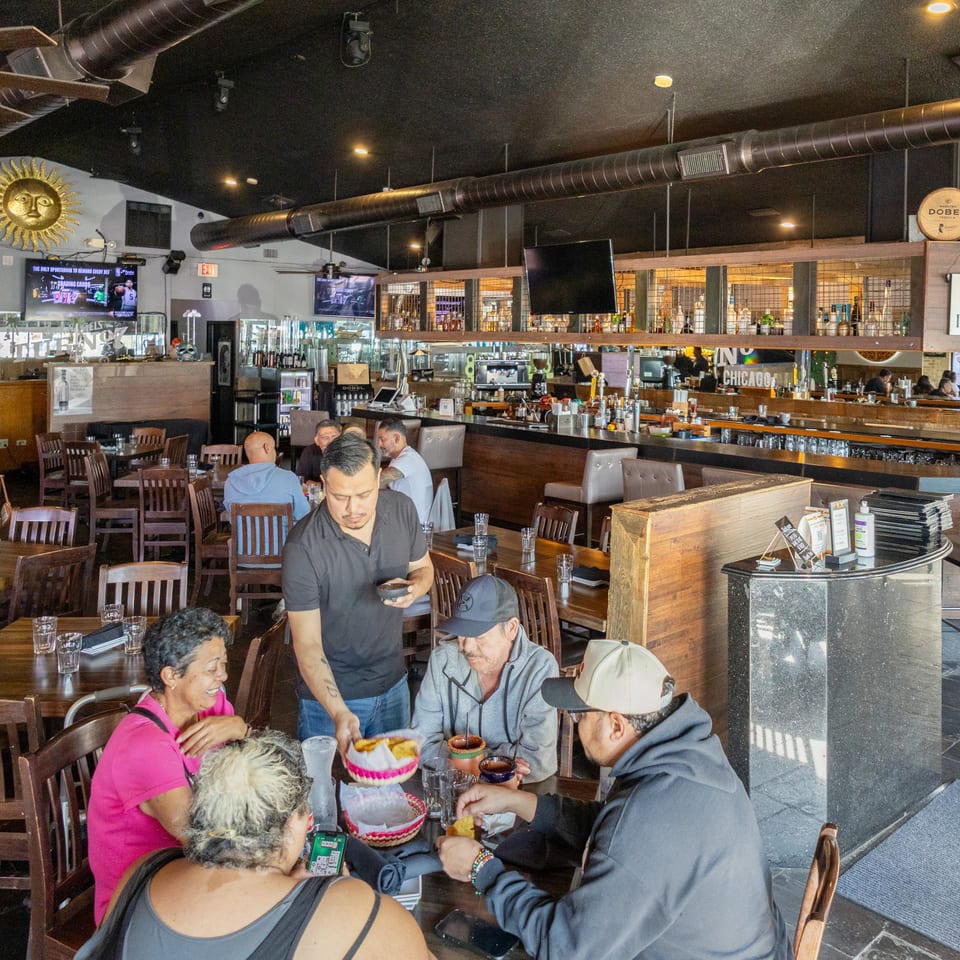 Lively Mexican restaurant interior with diners at tables, a long bar, exposed ductwork, televisions, and Corona Premier signage.