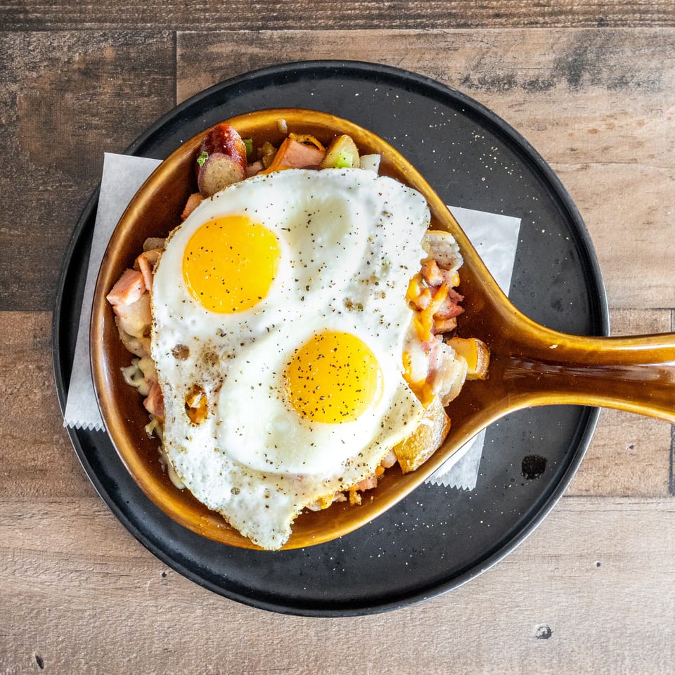 Skillet of potatoes, vegetables, bacon, and melted cheese topped with two sunny-side-up eggs on a black plate.