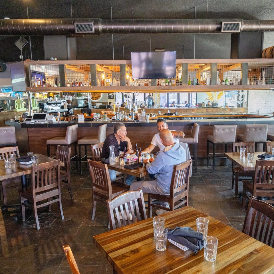Spacious restaurant dining room with wooden tables, bar seating, exposed ductwork, and guests dining beneath a television.