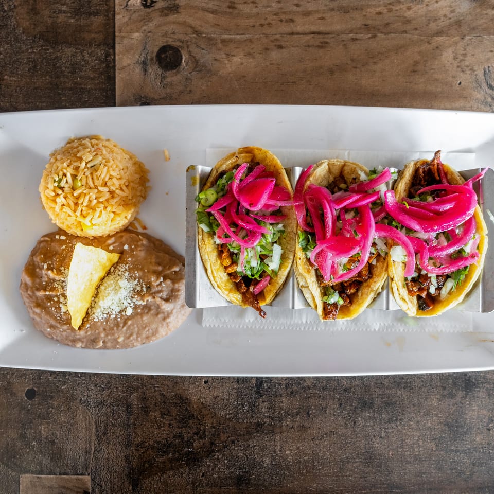 Three tacos topped with pickled onions, Mexican rice, and refried beans with cheese served on a white rectangular plate.