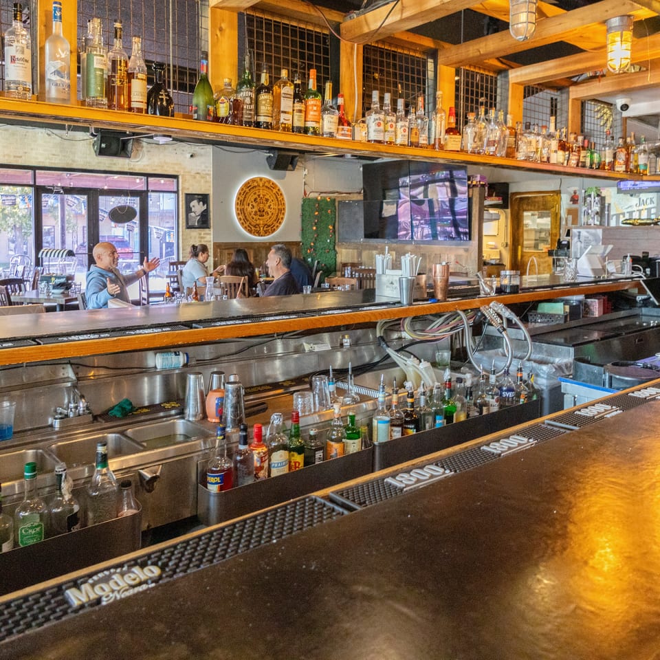 Busy restaurant bar stocked with liquor bottles, taps, glassware, televisions, and patrons seated near the windows.