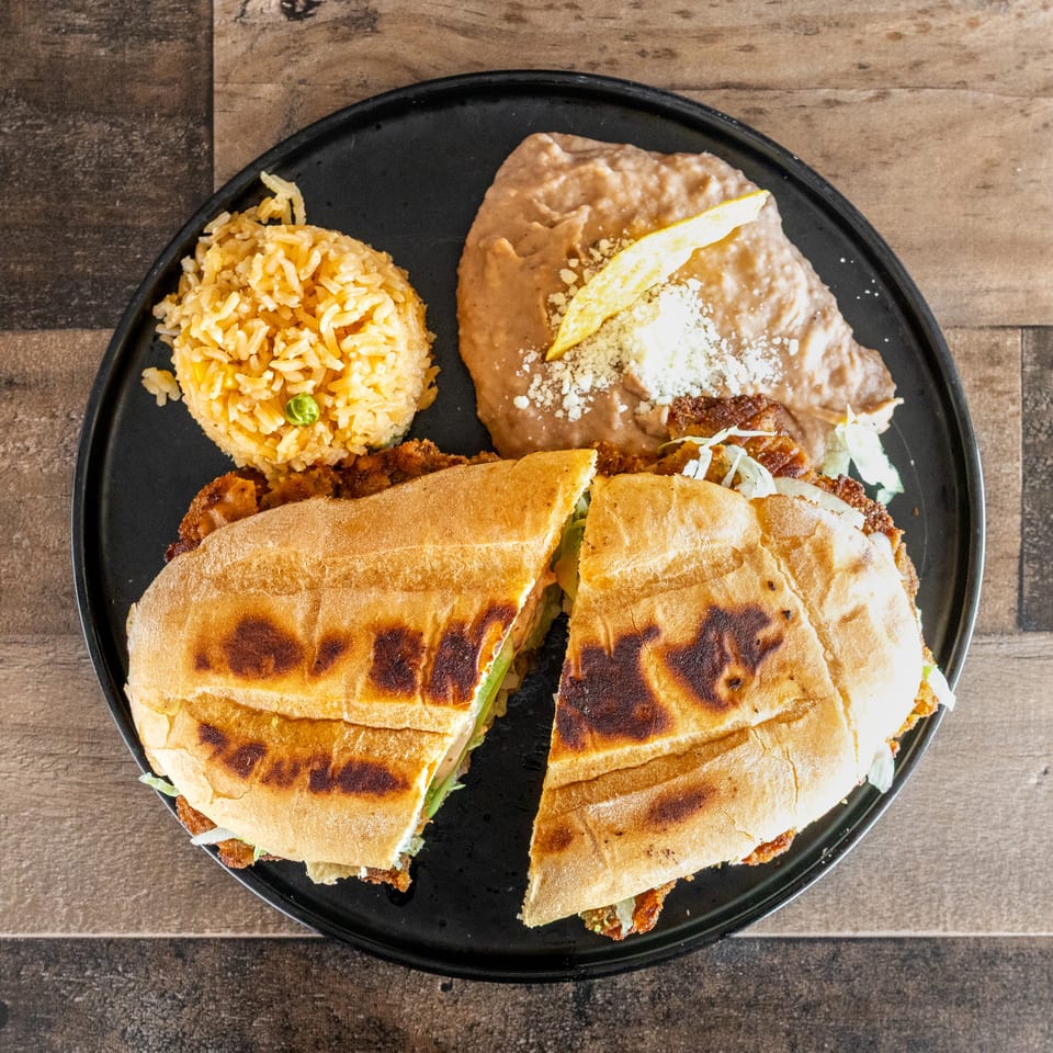 Black plate holding two toasted sandwiches, Mexican rice, and refried beans topped with cheese and a tortilla chip.